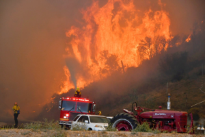 Firemen standing standing in a field spraying water on a giant wildfire.